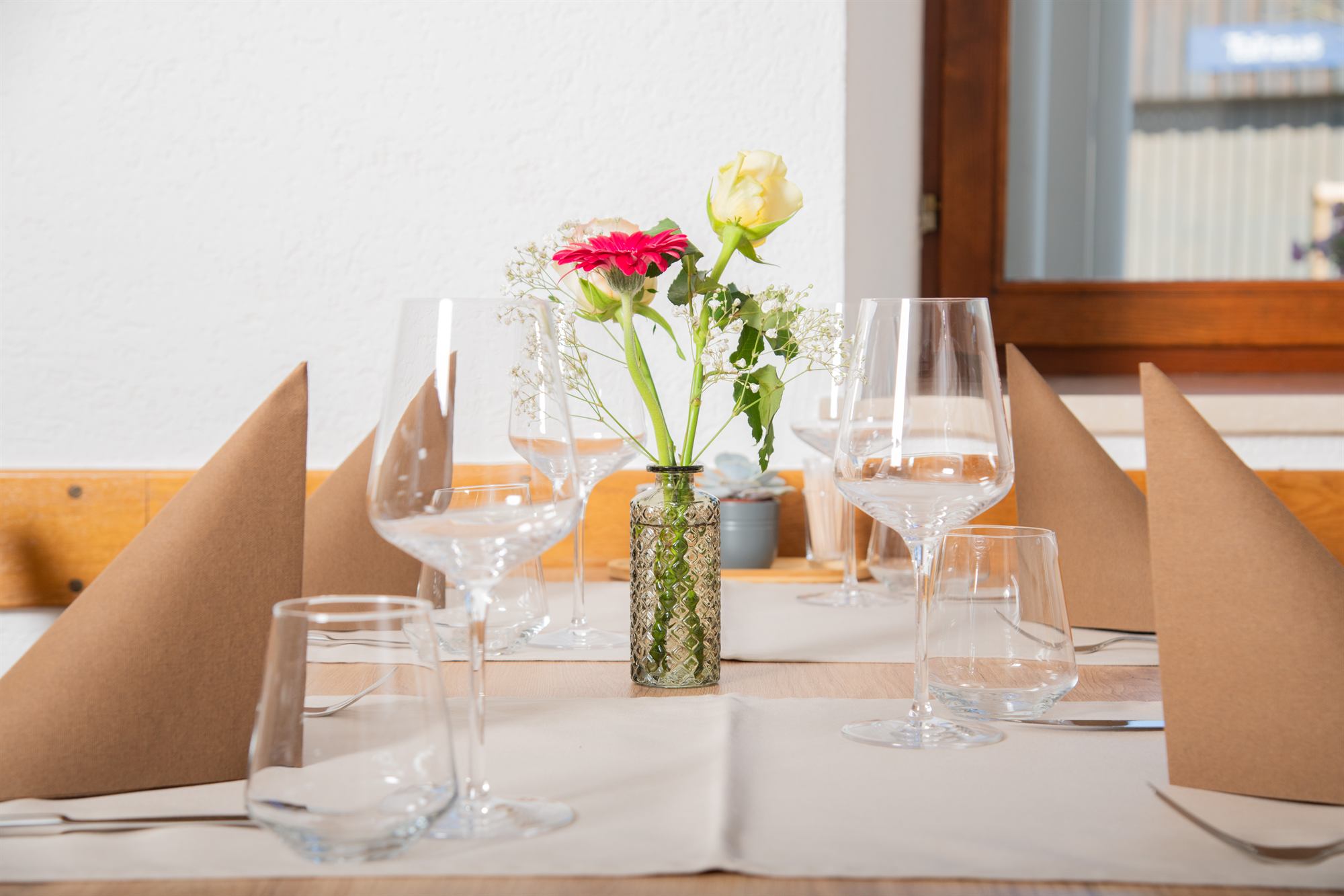 Liebevoll gedeckter Tisch mit Blumen und Weingläsern am Fenster.